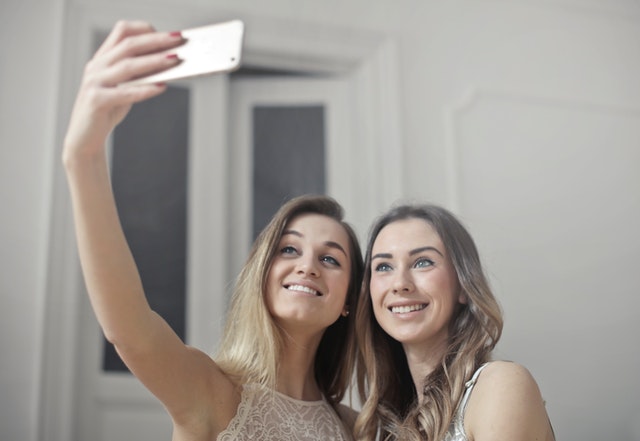 dvě světlovlasé ženy si dělají selfi fotku, jedna drží mobil v ruce, usmívají se na telefon